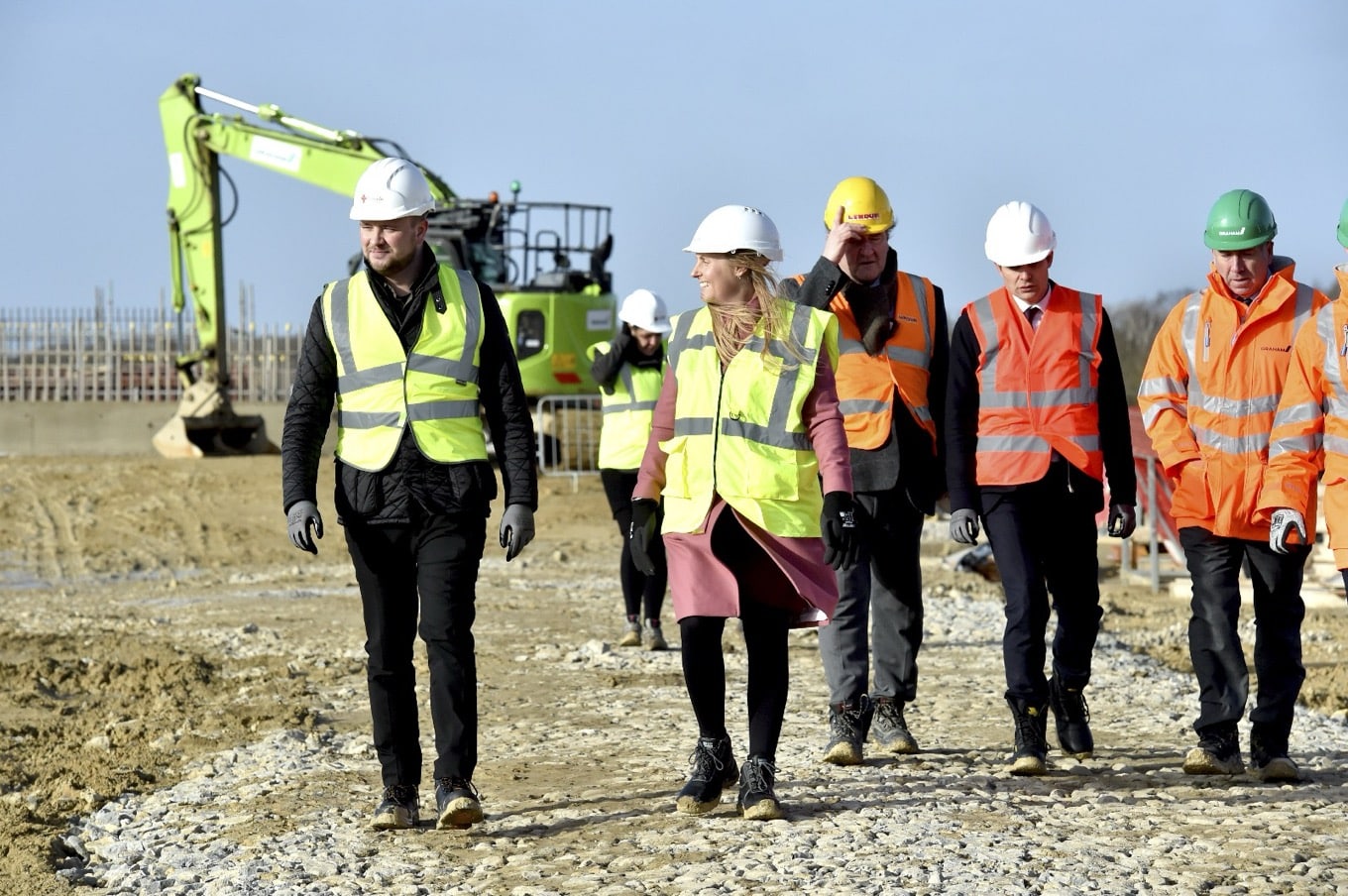A group of five people walking towards the camera on a building site in hi-vis jackets and a digger in the background.