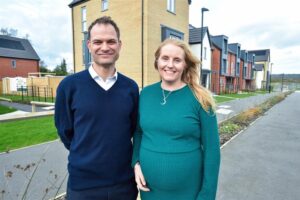 A white man and woman dressed smartly smiling for a posed photograph in front of new build houses.